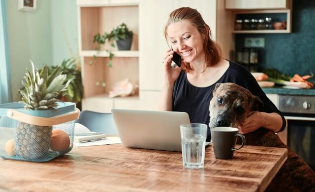 Woman on the phone with a dog in her lap. Both dog and woman are sitting at a table looking at an open laptop computer.